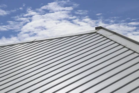Shiny silver metal roof panels on a house under a blue sky with scattered white clouds.