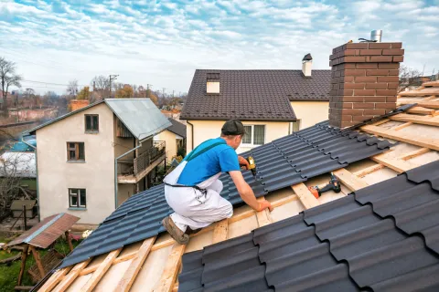 Worker installing dark gray metal roofing panels on a residential house roof on a clear day.