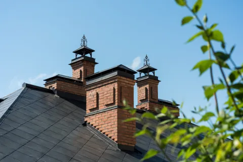 Close-up of a house roof with three brick chimneys against a clear blue sky and green foliage in foreground