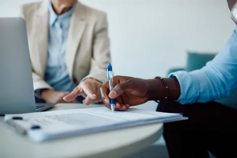 Two professionals reviewing and signing documents during a business meeting with a laptop on the table