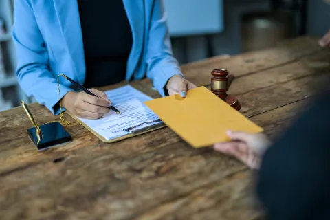 Person in blue jacket signing a legal document while handing an envelope across a wooden table with a judge's gavel nearby.