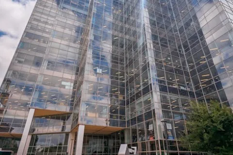 Modern glass office building with reflections, greenery, and clear sky above at the entrance plaza.