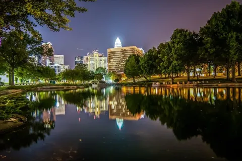 City skyline at night with illuminated buildings reflecting on calm river bordered by trees and park.
