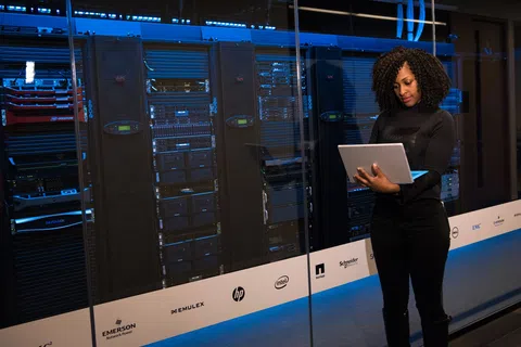 Woman using laptop in front of server racks in a data center with blue lighting.