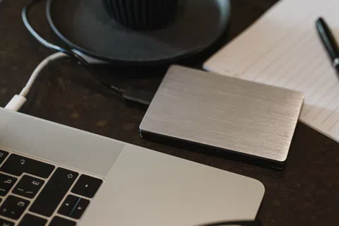 Silver external hard drive connected to a laptop on a dark desk near notebook and pen.