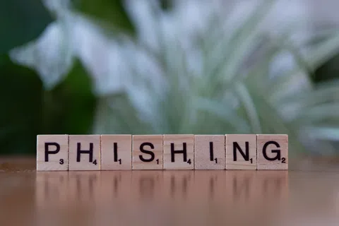 Wooden Scrabble tiles spelling phishing on a reflective surface with blurred green foliage background.