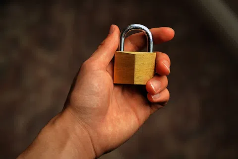 Hand holding a small brass padlock with a blurred dark background, symbolizing security and protection.