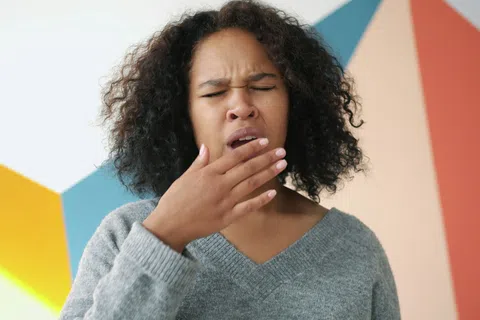 Woman with curly hair in gray sweater yawning and covering mouth against colorful geometric background