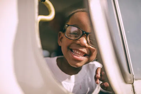 Smiling child wearing glasses looks through a car window on a sunny day, showing joy and innocence.