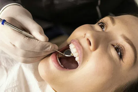 Dentist wearing gloves examines smiling woman's teeth with dental mirror during checkup.