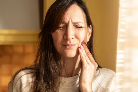 Woman in cream sweater holding her cheek with pain in a sunlit kitchen background