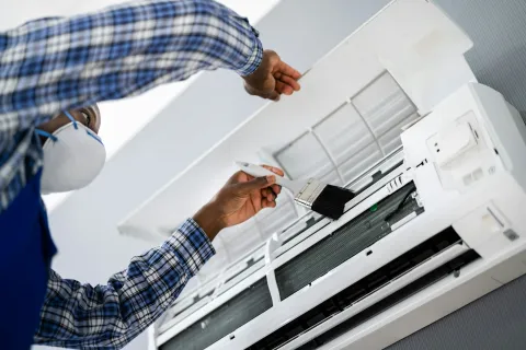 Technician cleaning an air conditioner unit with a brush while wearing a mask.