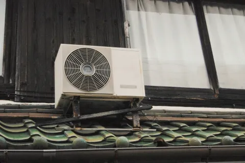 Air conditioning unit mounted on a building with traditional tile roof and wooden structure in the background.