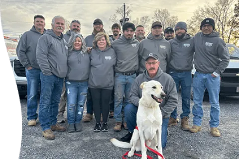 Team photo featuring employees in gray hoodies, with a dog in front, standing near work vehicles.