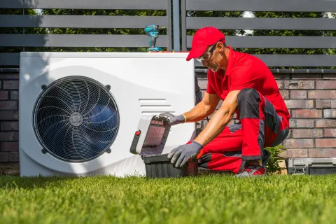 Technician in red uniform servicing outdoor HVAC unit on green lawn.