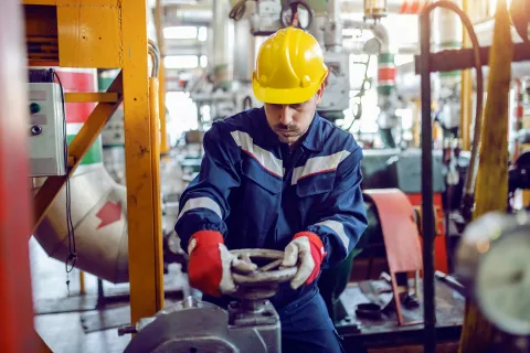 Industrial worker in safety gear adjusting valve in a factory with machinery and pipes around him