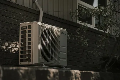 Outdoor air conditioning unit mounted on a brick wall with surrounding foliage and shadows.