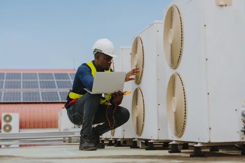 Technician in safety gear inspecting large HVAC units with a laptop and electrical testing tools outdoors on a rooftop.