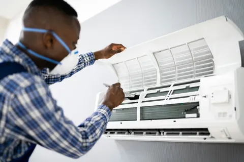 Technician wearing mask cleaning indoor air conditioner filters with brush for maintenance.