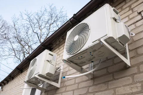 Two white outdoor air conditioning units mounted on a brick wall under a gutter with bare trees in the background.