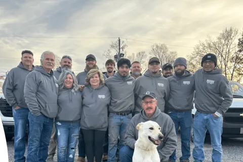 Group of people wearing gray Moody Electrical hoodies posing outdoors with a white dog on a cloudy day