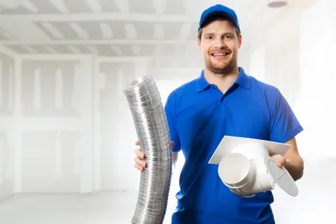 A smiling technician holds ductwork and a vent in a construction space, showcasing HVAC installation tools.