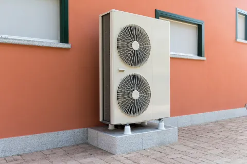Outdoor heat pump unit installed beside an orange wall with windows.