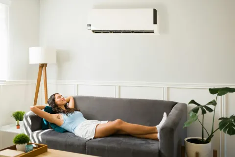 Woman relaxing on gray sofa under wall-mounted air conditioner in bright living room with plants and lamp
