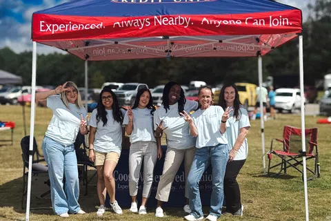 Six diverse women in white shirts smiling and posing under a Coosa Valley Credit Union tent outdoors on a sunny day.