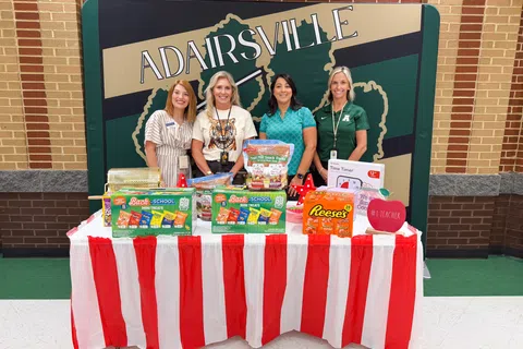 Four women standing behind a red and white striped table with school supplies and snacks at Adairsville school event.