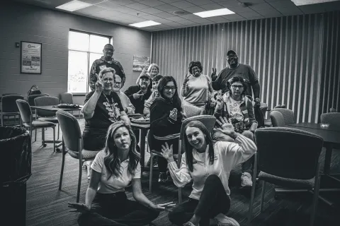 Black and white photo of diverse group of people smiling and posing playfully in a casual meeting room.