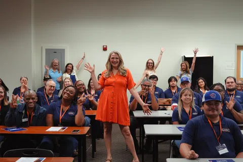 Group of diverse adults smiling and waving in a classroom with a woman in an orange dress standing at the center