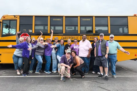 Group of cheerful adults posing in front of a yellow Cartersville City School bus in a parking lot.