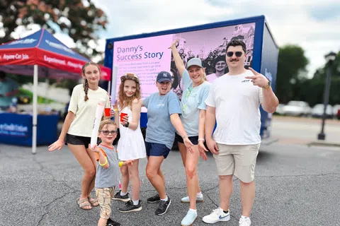 Group of smiling people posing outdoors near a colorful event display with casual summer clothing and lively expressions.
