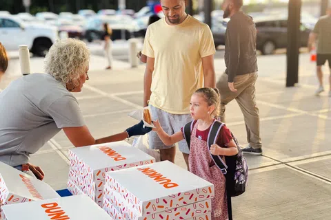 Young girl with backpack receives a Dunkin' donut from a vendor at an outdoor event with her father nearby.