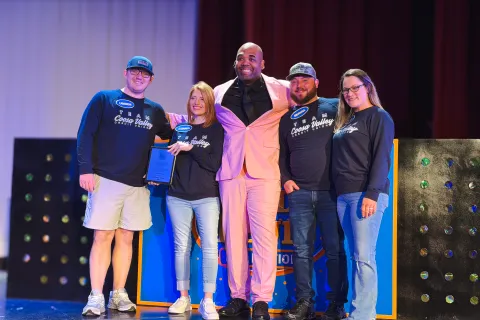 Five smiling people on stage, two holding a plaque, with one person in a pink suit and others in casual attire.