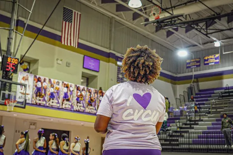 Woman wearing I love CVCU shirt watching cheerleaders in purple gymnasium with basketball hoop and American flag.
