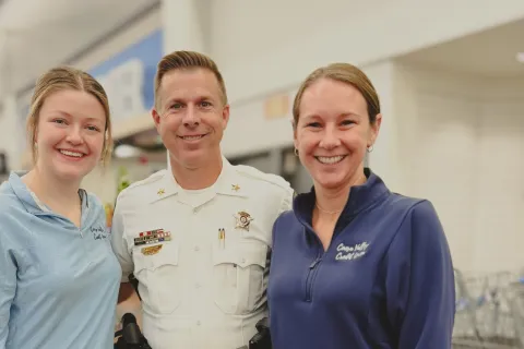 Two women smiling with a police officer in uniform inside a store aisle with shopping carts in the background.