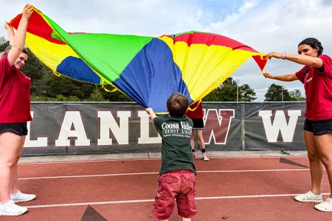 Children and adults playing with a colorful parachute on a running track in an outdoor sports area.
