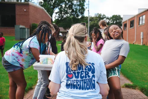 Children and an adult with kindness-themed shirt engage joyfully in outdoor activities at school playground.
