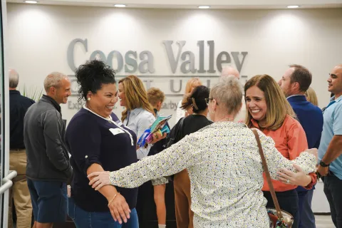 Smiling diverse group warmly greeting and embracing at Coosa Valley Community event in indoor setting.