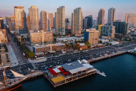 Aerial view of a modern waterfront city with high-rise buildings, pier restaurants, and calm ocean waters at sunset.