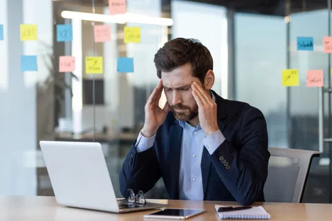 Stressed businessman in suit sitting at desk with laptop and sticky notes on glass wall in office.