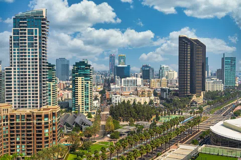 Downtown cityscape with modern high-rise buildings, palm trees lining the streets, and blue sky with clouds.