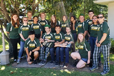 Group of smiling students wearing matching 4736 Patriots green shirts posing outdoors under trees.