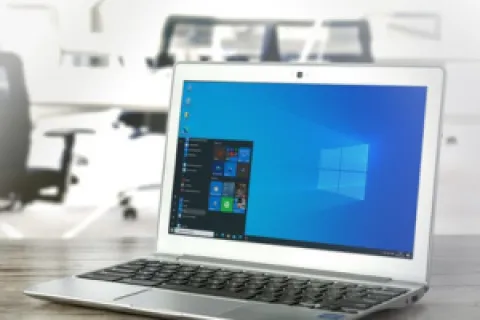Silver laptop on wooden desk displaying Windows 10 start menu in a bright modern office space