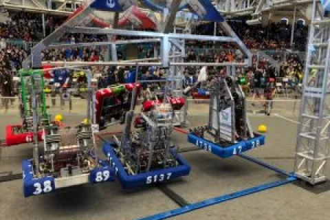 Three competition robots suspended mid-air during a robotics event with an audience in the background