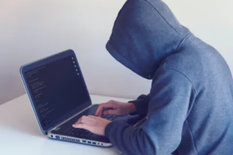 Person in gray hoodie typing code on laptop in a minimalistic white room, side view.