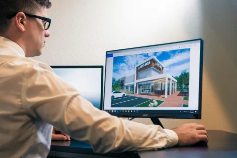 Man in glasses working on computer with architectural design of a modern commercial building on screen.