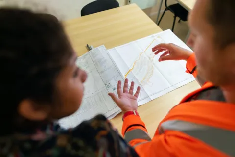 Two professionals reviewing construction plans on a wooden table, one wearing a high-visibility jacket.